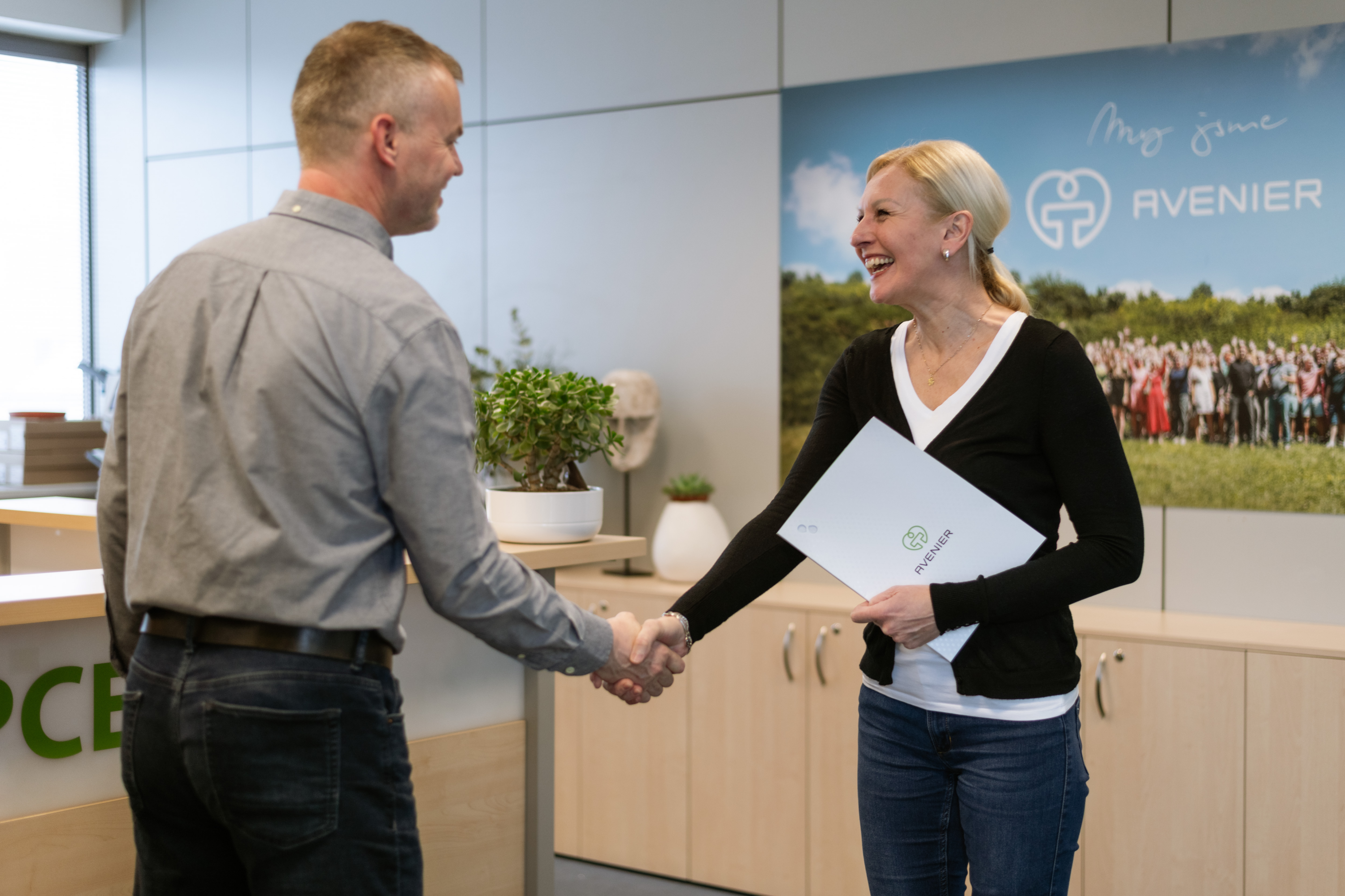 Man and woman shake hands in a bright office; the woman holds an AVENIER folder.**(Note: avoid markdown)