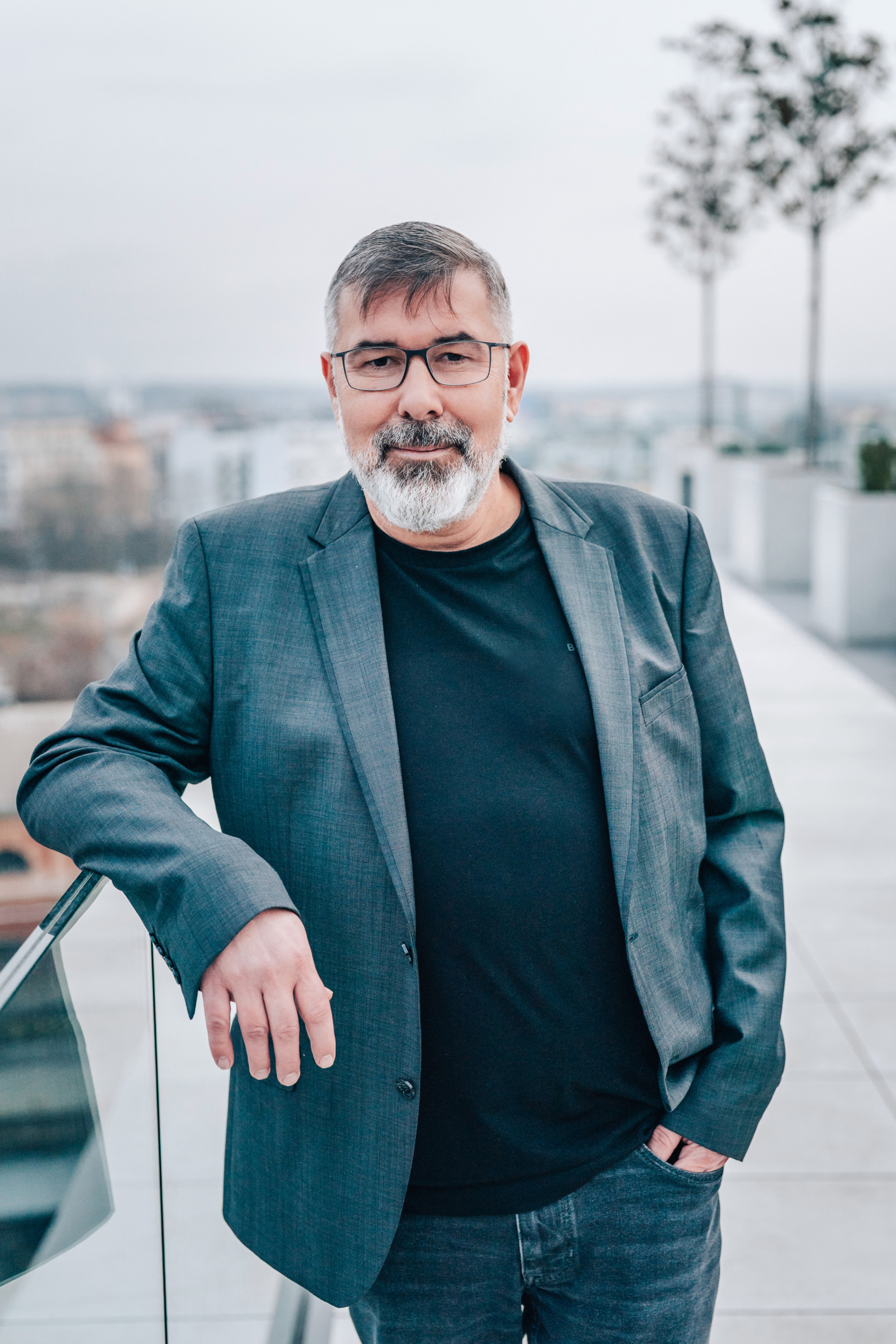 Man with gray beard and glasses in a blue blazer leaning on a glass railing with a cityscape in the background
